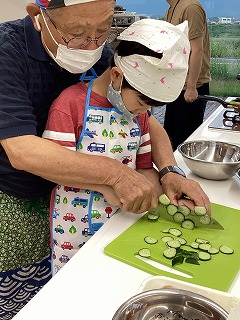おじいちゃんに手を添えられてキュウリをカットする子どもさん おじいちゃんに手を添えられてキュウリをカットする子どもさん